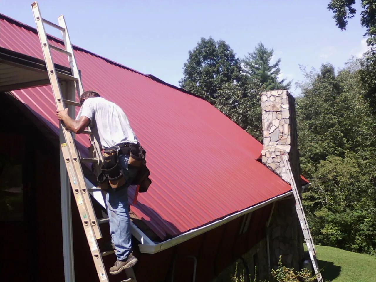 Roofer on a ladder beside a red metal roof and stone chimney — Soffit & Fascia Repair by Roofing Brothers in Butler